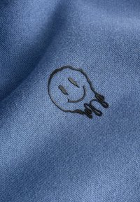 Visage souriant noir avec des gouttes brodées sur un tissu bleu texturé, créant un détail de conception ludique et décontracté.