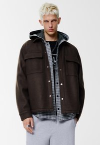 Giacca di lana marrone con cappuccio, camicia grigia a quadri sotto e pantaloni della tuta grigi. Presenta bottoni a pressione e doppie tasche frontali.