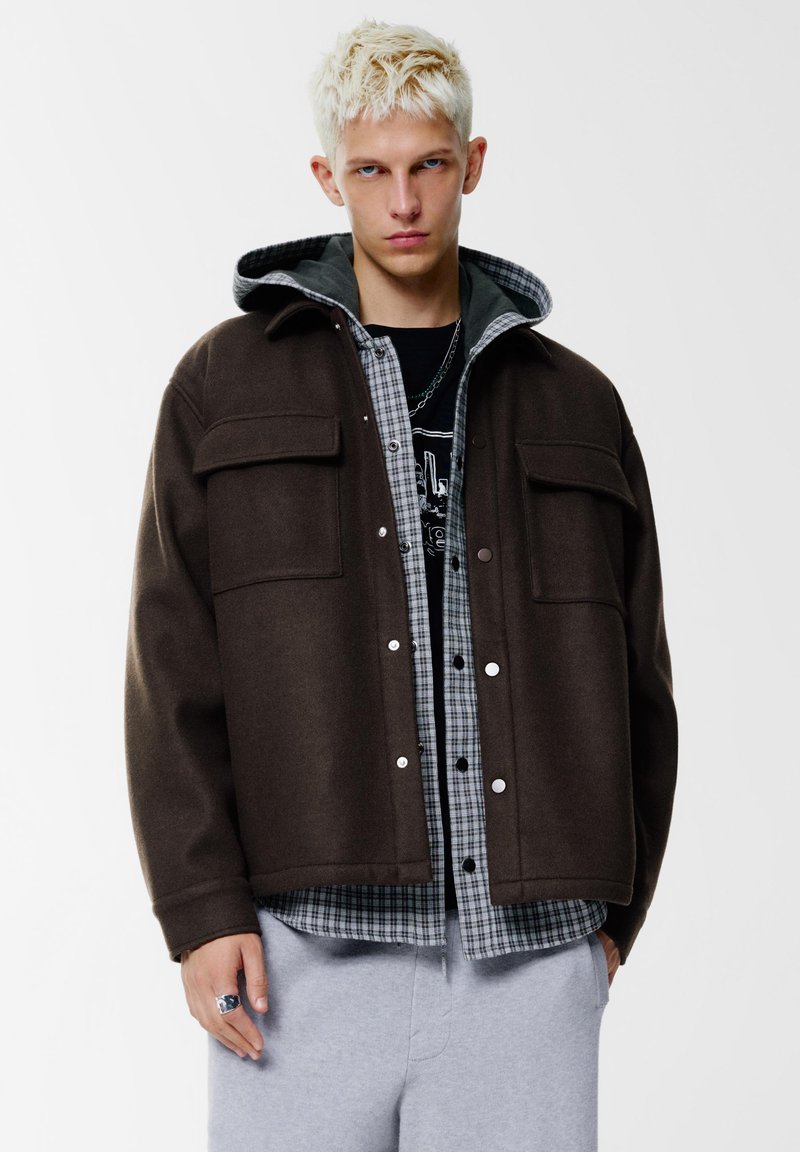 Giacca di lana marrone con cappuccio, camicia grigia a quadri sotto e pantaloni della tuta grigi. Presenta bottoni a pressione e doppie tasche frontali.