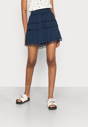 Woman wearing navy blue tiered mini skirt with lace trim, white sleeveless top, and white buckle sandals standing on light floor.