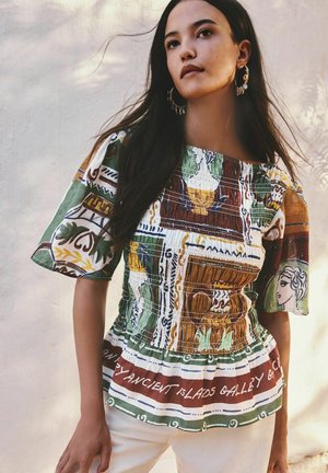 Frau mit langen dunklen Haaren, die ein gemustertes, gerafftes Oberteil mit kurzen Ärmeln und große Creolen trägt, steht vor einer hellen Wand.