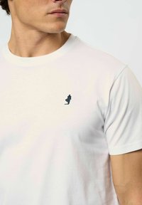 Vīrietis valkā vienkāršu baltu T-kreklu ar apaļu kakla izgriezumu un mazu melnu logotipu ar staigājoša cilvēka attēlu kreisajā krūtīs.