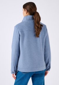 Chaqueta de mezcla de lana azul con un alto cuello, hombros caídos y puños abotonados. Textura suave con un diseño limpio y minimalista.