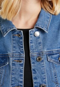 Jeansjacka i ljusblått med silverfärgade metallknappar, bröstfickor och kontrasterande sömmar. Tyget har en något sliten textur.