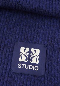 Tissu bleu foncé texturé avec des côtes verticales, présentant une étiquette carrée avec des graphismes blancs stylisés et le mot « STUDIO ».