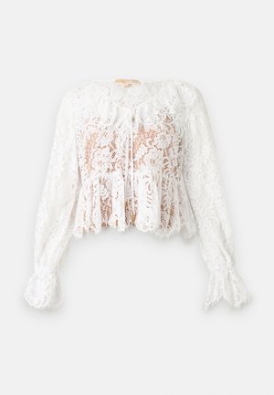 Blouse blanche à manches longues en dentelle florale avec ourlet péplum, fermeture à nouer devant et poignets festonnés sur un fond uni.
