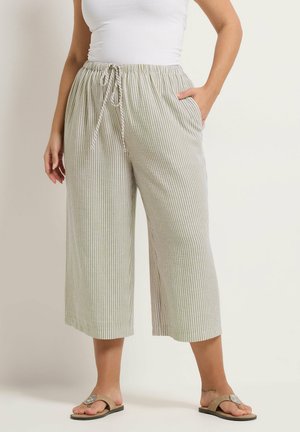 Kvinde iført hvid tanktop og beige, brede, stribede bukser med snøre i taljen, stående med den ene hånd i lommen og beige sandaler.