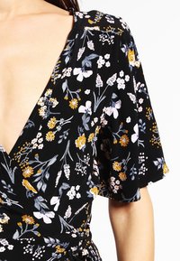 Tejido negro con estampado floral en amarillo, blanco y gris en un vestido envolvente con escote en V profundo y mangas cortas con volante.