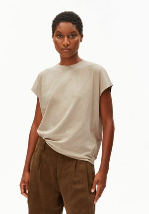 Vrouw met kort krullend haar die een beige T-shirt met korte mouwen draagt, ingestopt in een hooggetailleerde bruine broek, staand tegen een witte achtergrond.