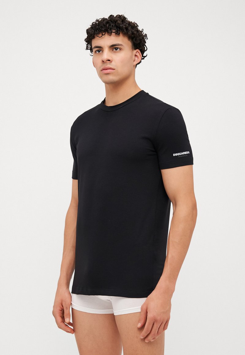 Schwarzes Baumwoll-T-Shirt mit Rundhalsausschnitt und kurzen Ärmeln. Mit einem weißen Logo auf dem linken Ärmel. Glatte Textur, taillierte Passform.