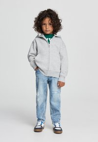 Felpa grigia con zip e colletto alto, indossata su una maglietta verde, abbinata a jeans azzurri chiari strappati e sneakers blu navy.