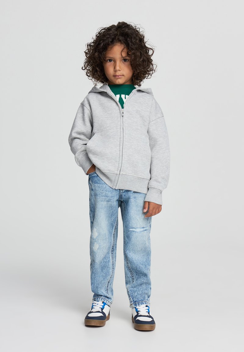 Felpa grigia con zip e colletto alto, indossata su una maglietta verde, abbinata a jeans azzurri chiari strappati e sneakers blu navy.