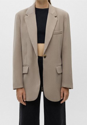 Persoon draagt een beige oversized blazer over een zwart cropped geribd topje en zwarte high-waisted broek, staand voor een lichte achtergrond.