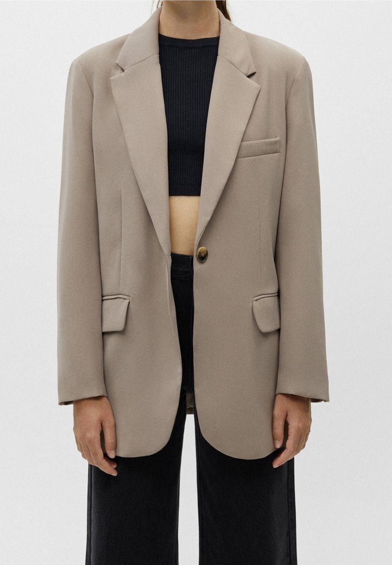 Personne portant un blazer beige oversize par-dessus un top court côtelé noir et un pantalon noir taille haute, debout devant un fond clair.