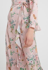 Robe portefeuille fleurie en tissu rose doux, avec des manches à volants, une ceinture nouée à la taille, et un mélange de motifs floraux colorés sur toute la surface.