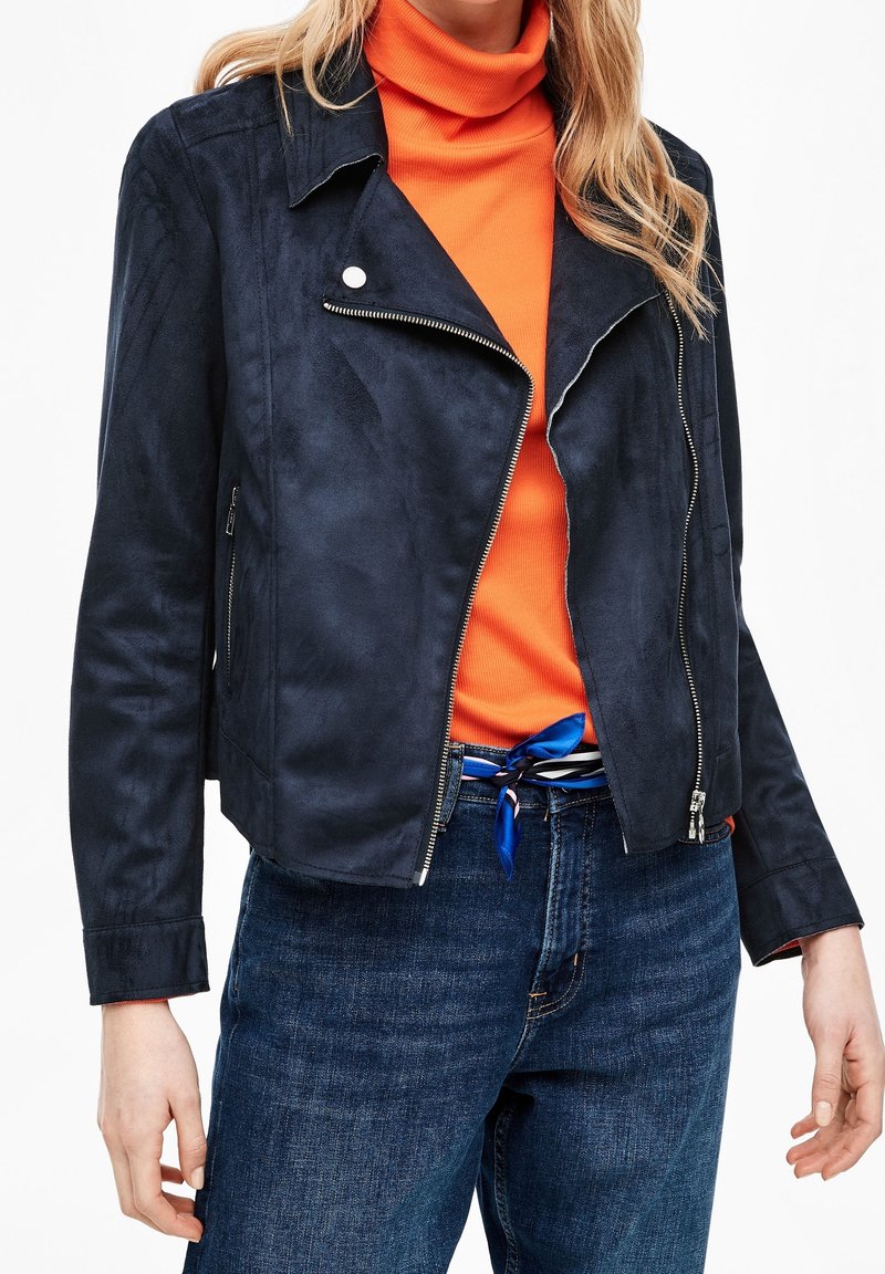Giacca da moto in suede blu navy con minuterie in argento, con zip frontale e colletto a punta, indossata sopra un dolcevita arancione e jeans blu.