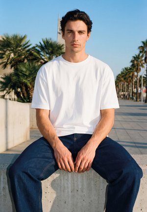 Mladi muškarac s tamnom kosom, u bijeloj majici i plavim trapericama, sjedi na betonskoj lajsni s palmama i bistrim nebom u pozadini.