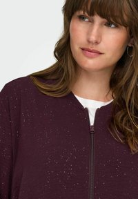 Bordo bomber jakna na zatvaranje sa teksturiranom tkaninom i rasutim srebrnim šljokicama. Ima okrugli izrez i rebrasti detalj.