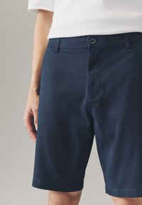 Tumesinised lühikesed püksid, mis on valmistatud silesest kangast, varustatud nööpkinnitusega, esipõllega ja sirge lõikega, ulatudes veidi kõrvale põlvest.