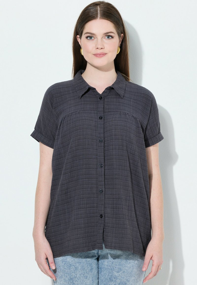 Camisa de botones de manga corta en gris oscuro con una tela texturizada. Cuenta con un cuello con solapa y un bolsillo frontal. Corte relajado con un dobladillo curvado.