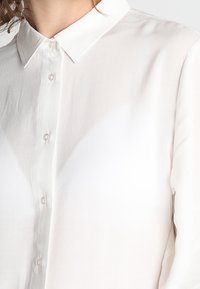 Camicia bianca abbottonata con colletto classico, texture liscia e bottoni lucidi, presenta un aspetto leggermente trasparente.