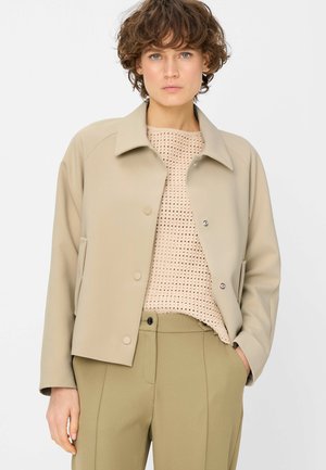 Woman with short curly hair wearing a beige buttoned jacket, light knitted sweater, and olive-green pants with one hand in pocket.