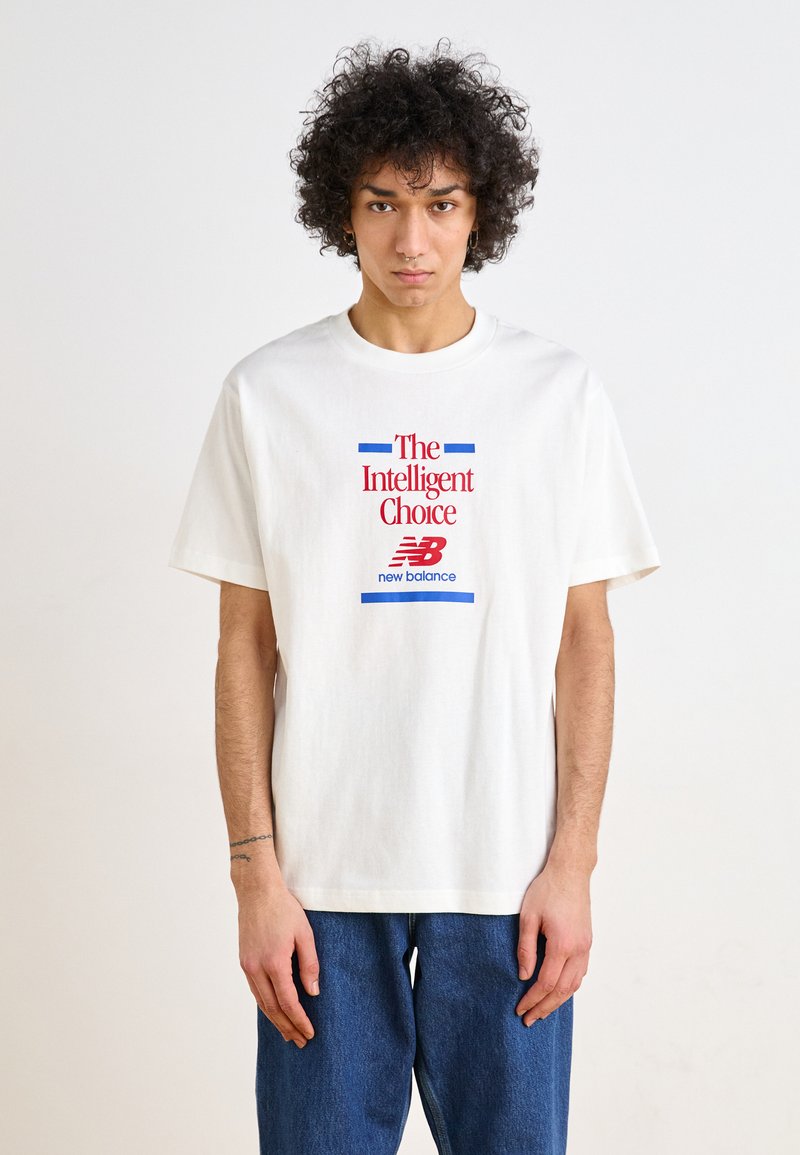 T-shirt de algodão branco com mangas curtas, apresentando um design gráfico em vermelho e azul que diz "A Escolha Inteligente" e "new balance."