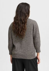 Pull en tricot marron avec une coupe ample, texture côtelée, col rond et manches trois-quarts. Porté avec un pantalon noir ample.