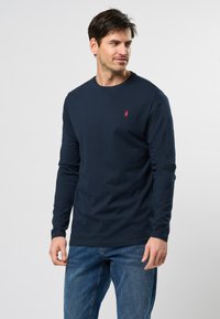 Uomo che indossa una camicia a maniche lunghe blu navy con un logo rosso sul petto e jeans blu, mentre guarda a destra contro uno sfondo neutro.
