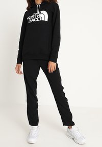 Felpa nera con logo bianco, cappuccio con cordoncino e tasca a marsupio, abbinata a pantaloni neri con strisce laterali e sneakers bianche.