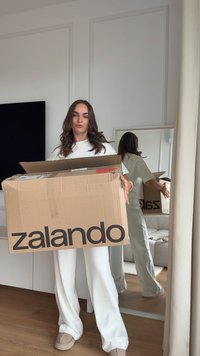 Ein Karton mit dem Zalando-Logo in fettem Schwarz, gehalten von einer Person in heller Kleidung. Im Hintergrund sind ein Spiegel und ein Sofa zu sehen.