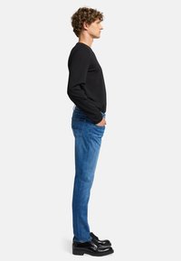 Maglietta nera a maniche lunghe, jeans blu slim-fit e scarpe di pelle nere. I jeans presentano dettagli di cucitura nelle tasche posteriori. Vista laterale del modello.