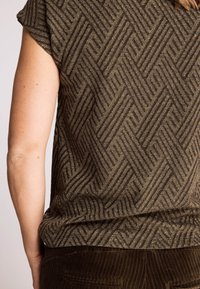 Kurzarm-Oberteil aus olivgrünem Stoff mit Fischgrätmuster und metallischen Fadenakzenten. Strukturierte Oberfläche mit lockerem Schnitt.