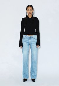 Femme debout devant un fond uni, portant un haut noir à manches longues, un jean bleu clair, des chaussures noires et les cheveux en deux tresses.