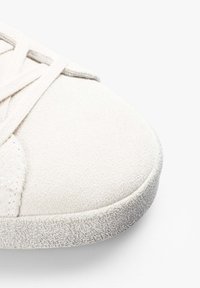 Zapatilla blanca de ante con suela de goma texturizada, punta redondeada y patrón de cordones cruzados. Acabado suave con diseño minimalista.