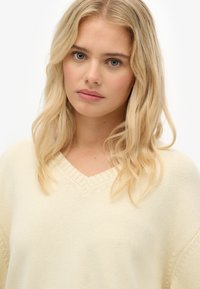 Blondynka z falującymi włosami, nosząca kremowy sweter z dekoltem w serek, patrząca prosto przed siebie z neutralnym wyrazem twarzy na białym tle.
