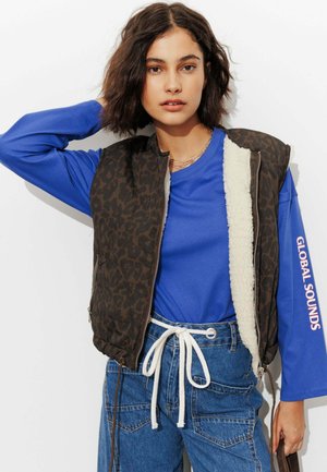 Jeune femme aux cheveux bruns ondulés porte une chemise bleue à manches longues avec le texte "GLOBAL SOUNDS", un gilet à motif léopard et un jean taille haute avec un cordon blanc.