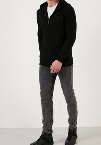 Felpa nera con zip, in tessuto testurizzato, dotata di tasche frontali e cappuccio con cordini, abbinata a jeans grigi e sneakers nere.