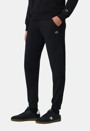 Persona con pantalones deportivos negros y sudadera a juego con logo, zapatillas negras con detalles en blanco, de pie con una mano en el bolsillo.