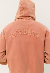 Personne portant un sweat à capuche rose délavé avec le texte brodé "LES DEUX" sur le haut du dos, debout dos à la caméra.
