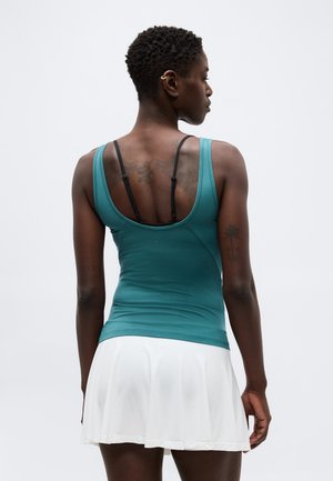 Γυναίκα με κοντά σγουρά μαλλιά που φοράει τιρκουάζ μπλούζα χωρίς μανίκια και λευκή φούστα, εμφανιζόμενη από πίσω με εμφανή τατουάζ στα χέρια και στο πάνω μέρος της πλάτης.