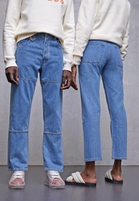Světle modré džínové kalhoty s volnějším střihem, zakončené panely a mírně třepenými lemy, doplněné bílou obuví.
