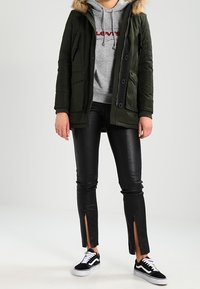 Oliwkowo-zielona parka z kapturem podszytym futrem, pod spodem szara bluza z kapturem, czarne skórzane spodnie z rozcięciami na końcach nogawek oraz czarno-białe sneakersy.