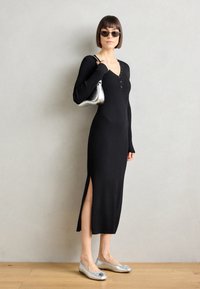 Abito midi nero a costine con maniche lunghe, spacco laterale e dettagli in bottone sul collo. Abbinato a ballerine argentate e una borsa metallica.