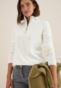 Pull blanc en tissu texturé avec un col à demi-zip, associé à un jean gris foncé. La personne tient une veste olive verte pliée.