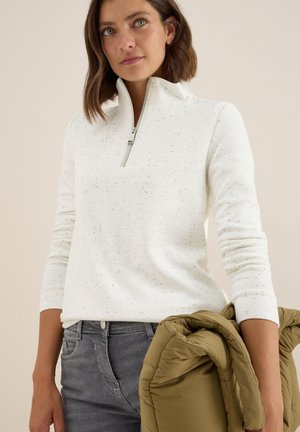 Weißer Pullover aus strukturiertem Stoff mit einem halbseitigen Reißverschlusskragen, kombiniert mit dunkelgrauen Jeans. Die Person hält eine gefaltete olivgrüne Jacke.