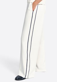 Pantaloni bianchi a gamba larga realizzati in tessuto morbido, con righe laterali color navy e una tasca sul lato destro. Footwear strutturato di un blu scuro completa il look.