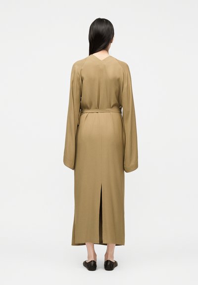 Femme aux longs cheveux foncés portant une robe beige ceinturée à manches longues et longueur cheville, ainsi que des chaussures plates noires, dos tourné sur un fond uni.