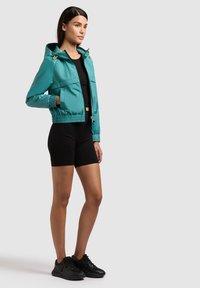 Žena stojaca v tyrkysovej mikine s kapucňou na zips, čiernom krátkom topiku, čiernych cyklistických kraťasoch a čiernych teniskách na sivom pozadí.