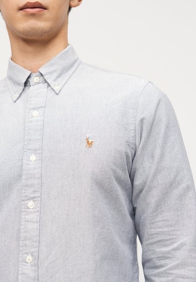 Άνδρας που φοράει ανοιχτό μπλε πουκάμισο με κουμπιά και μικρό κεντημένο λογότυπο παίκτη πόλο πάνω σε άλογο στο στήθος.